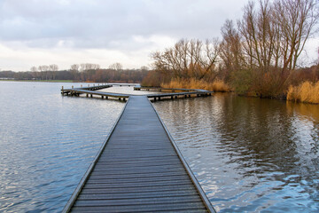 Obraz premium Panoramic view of the shore of Kralingen Lake, Rotterdam, the Netherlands during winter with long stretched boardwalk in the water and lake lined with leafless trees