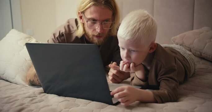A blond man in glasses with a beard, together with his little albino son with blue eyes in a brown jacket, watch a movie on a laptop and educational cartoons during their relaxation and spending time