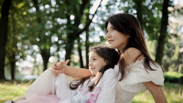 A daughter and her mother are sitting on the grass in a park. The Concept of motherhood and happy childhood
