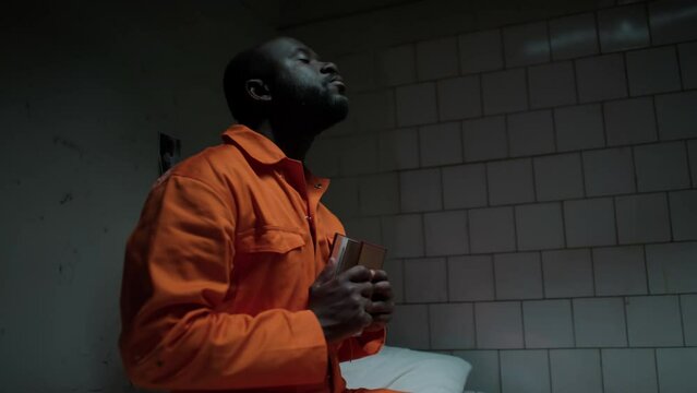 African American inmate sitting on bed in prison cell, holding bible close to chest and praying