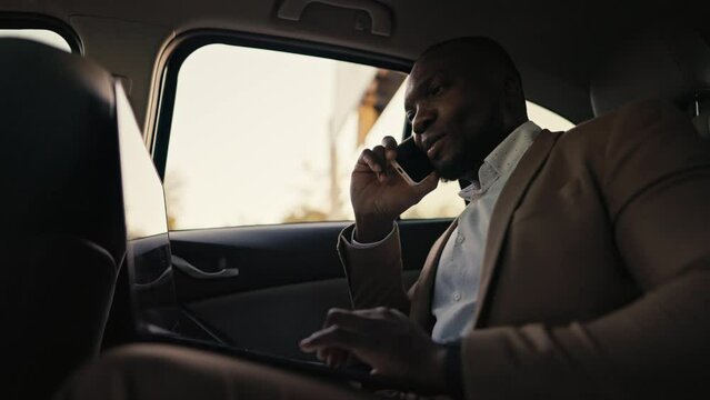 Happy and confident man Businessman with Black skin color in a brown jacket communicates on a smartphone and types on his laptop while driving In the passenger seat in a modern car outside the city
