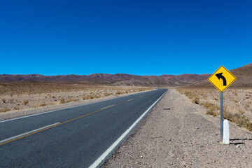 Yellow road sign with a black arrow pointing to the left (road goes to the left) along the road in the Andes mountains, Salta, Argentina, South America
