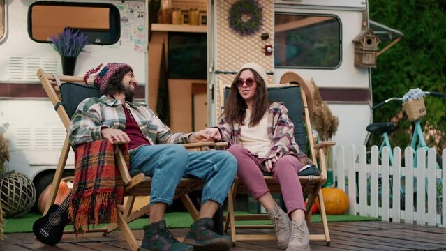 Happy brunette girl in round glasses and a white hat in a plaid shirt and pink pants sits on a chair with her brunette boyfriend in a hat near a trailer during their picnic at a camp outside the city