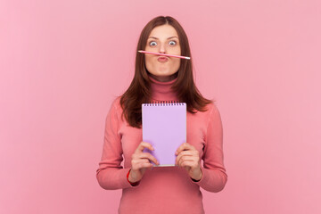 Funny optimistic woman with brown hair showing empty paper of organizer, holding pen as mustache, having fun, wearing rose turtleneck. Indoor studio shot isolated on pink background