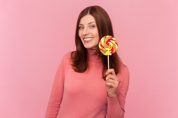 Sweet rainbow candy. Positive woman with brown hair holding lollipop and looking at camera with happy expression, wearing rose turtleneck. Indoor studio shot isolated on pink background