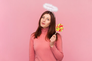 Winsome cute charming woman with brown hair and nimb over head, looking at camera and holding colorful delicious lollipop, wearing rose turtleneck. Indoor studio shot isolated on pink background - obrazy, fototapety, plakaty