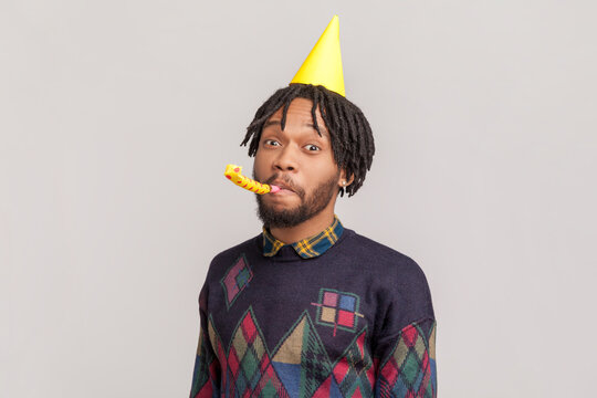 Portrait of positive funny african-american man in yellow festive cone with dreadlocks and beard blowing party horn, looking at camera. Indoor studio shot isolated on gray background.