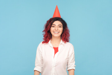 Portrait of festive happy woman with fancy red hair in party cone, celebrating birthday, smiling...