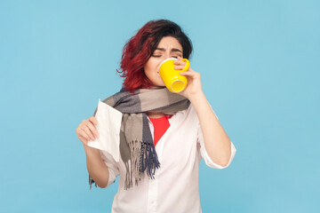 Portrait of sick unhealthy ill woman with fancy red hair has flu symptoms, drinking tea, suffering runny nose and high temperature, wearing white shirt. Indoor studio shot isolated on blue background