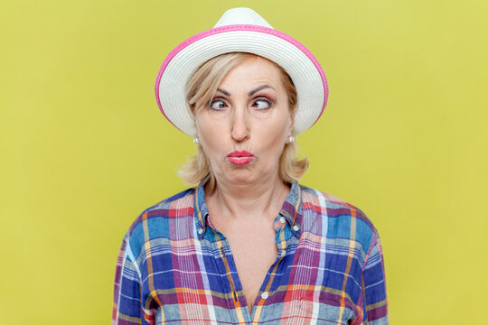 Portrait of funny foolish mature woman wearing checkered shirt and hat making grimace and crosses eyes, playing fool, having fun. Indoor studio shot isolated on yellow background. - Powered by Adobe