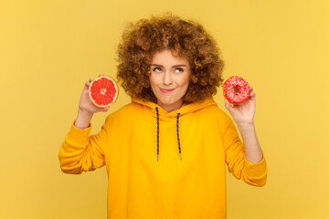 Portrait of hungry crazy woman with Afro hairstyle holding tasty doughnut and half of grapefruit,...