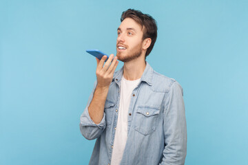 Portrait of unshaven man holding mobile phone in hand, giving commands to voice assistant, leaving voice message or recording audio notes, wearing denim shirt. Indoor shot isolated on blue background.