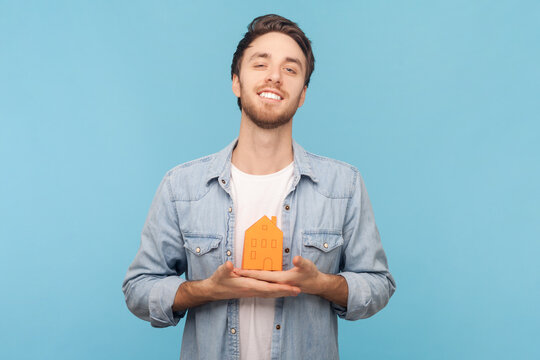 Cheerful handsome unshaven man holding in hands little paper house looking at camera with pleasant smile, dreaming about own house, wearing denim shirt. Indoor shot isolated on blue background.