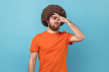Portrait of man with Afro hairstyle wearing orange T-shirt looking at camera and nose in disgust, avoiding unpleasant smell of farting, stinky aroma. Indoor studio shot isolated on blue background. - obrazy, fototapety, plakaty