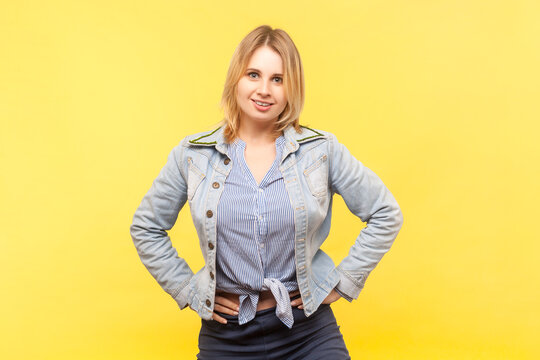 Portrait of good looking charming blonde woman wearing denim jacket standing with hands on hips looking at camera with satisfied expression. Indoor studio shot isolated on yellow background.