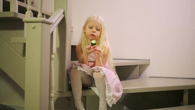 Girl in a pink dress is sitting on stairs and eating a cucumber