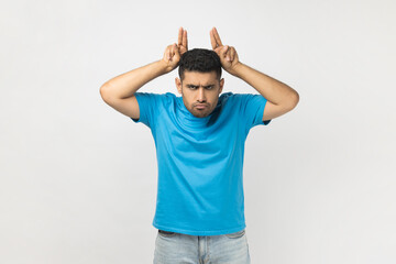 Portrait of angry strict handsome unshaven man wearing blue T- shirt standing showing bully gesture, looking at camera with aggression, attack. Indoor studio shot isolated on gray background.