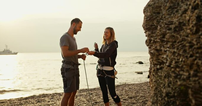 A blonde girl with curly hair in black clothes teaches her brunette boyfriend in a gray T-shirt to tie special knots in order to make belay while folding