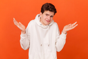 Portrait of clueless confused man wearing white hoodie shrugging shoulders as doesn't know answer, can`t make decision, being uncertain, not sure. Indoor studio shot isolated on orange background.