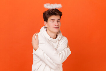 Portrait of young man with nimb above his head wearing white hoodie, embracing himself and smiling with pleasure, feeling self-pride. Indoor studio shot isolated on orange background.
