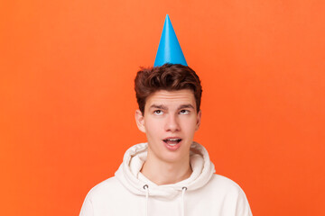 Portrait of amazed excited young man wearing white hoodie, standing with astonished expression, looking up at blue party cone on his head. Indoor studio shot isolated on orange background.