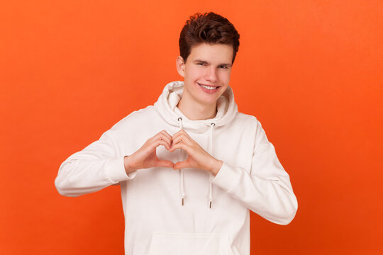 Portrait of pleased good looking man wearing white hoodie shapes heart over chest, confesses in love, has romantic mood, expresses truthful feelings. Indoor studio shot isolated on orange background.