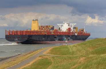a scenic coast landscape with a big container ship that sails very close to the green seawall at the dutch coast in zeeland and a beautiful sky