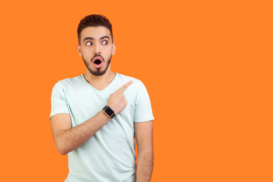 Portrait of astonished young bearded man wearing T-shirt standing with open mouth pointing away at advertisement area, copy space. Indoor studio shot isolated on orange background.