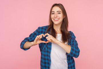 Portrait of romantic smiling joyful brown haired woman showing heart shape with hands in front of her chest, wearing checkered shirt. Indoor studio shot isolated on pink background.