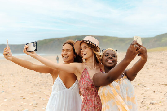 multiracial group of three friends on the beach taking pictures with their smartphones to show on social networks during a summer trip to the coast.