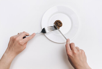 Two female hands hold a stainless steel knife and fork near the plate and cut artificial feces. Fake news, jokes. Concept of inept chef, lousy restaurant, bad food, crappy taste.