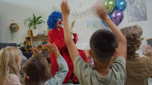 Back slowmo of unidentified happy kids watching clown exploding confetti shot tube at birthday party at home