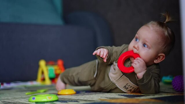 Infant child in a romper lies on the floor. Cute baby picks a toy and takes it to his mouth. Blurred backdrop.