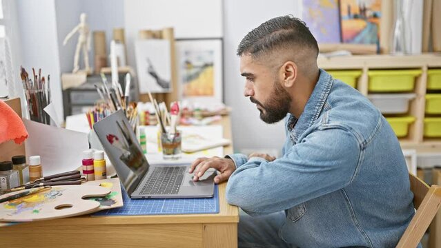 Bearded man in denim using laptop at artist studio with paintings and art supplies.