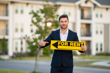 Successful real estate agent in a suit holding for rent sign near new apartment. Realtor or real estate agent shows board for rent. Rent home, rental concept. Renting an apartment.