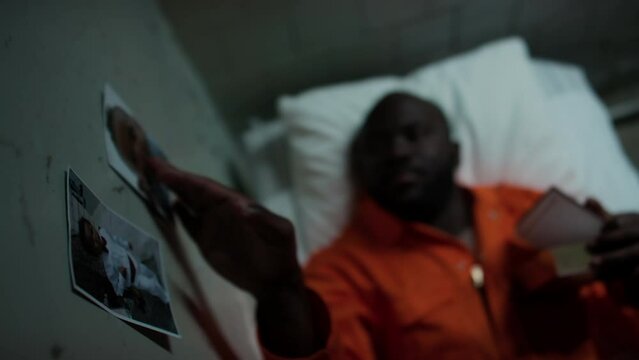 High angle view of Black inmate resting on bed in prison cell, reading bible and touching photos of beloved baby son