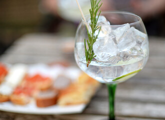 Glass of gin and tonic with ice cubes, rosemary and lime, plate with snacks on the side. Selective focus