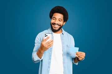 A young Latin man beams with a smile as he looks at his smartphone, holding a credit card in his...