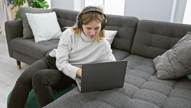 A focused woman with headphones uses a laptop on a sofa in a cozy living room, depicting work from home.