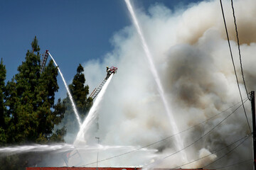 Firemen attacking a fire with water canon from the top of a hook and ladder fire engine with the ladder extended, lots of smoke, and a blue sky background.