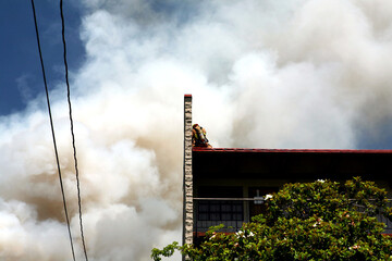 A firefighter taking shelter from a blazing fire and its smoke behind a wall on top of a roof.