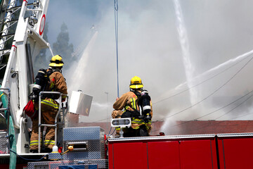 Fototapeta premium Firemen standing on a hook and ladder fire engine watching as water canons blast a structure fire with streams of water.