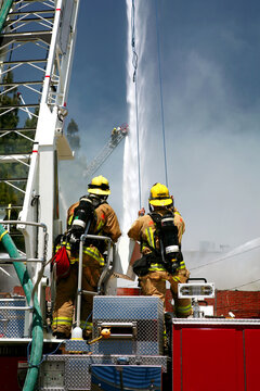 Vertical image of Firefighters standing on a hook and ladder fire engine watching as water canons blast a structure fire with streams of water.