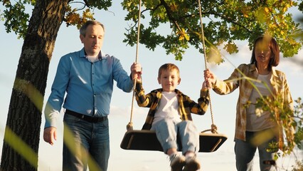 Caring grandma and grandpa push grandson on swing enjoying time together. Grandparents engaged in...