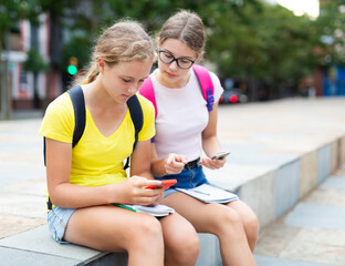 Two young girls sitting in park and doing homework. They're using smartphones to complete tasks.