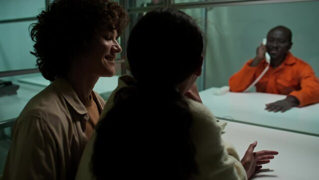 Rack focus shot of Black inmate sitting behind glass wall and having phone conversation with family visiting him in prison