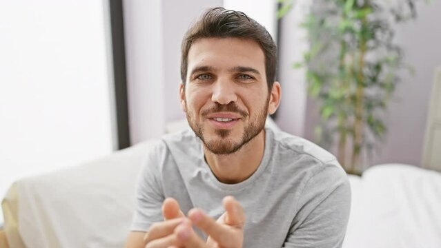 A young hispanic man with a beard smiles and gestures a kiss in a bedroom setting, portraying a casual and friendly demeanor.