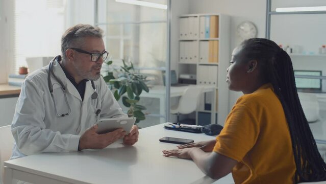 Medium shot of middle-aged male GP in white lab coat, with stethoscope sitting in clinic with overwhelmed black female patient just diagnosed with diabetes, explaining bloodwork results on tablet