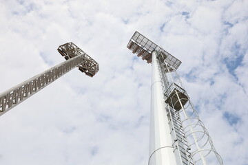 Metal poles of stadium lights. Closeup of new silver galvanized tall mast and steel frame tower of spotlights in bottom view on sky background with white clouds with selective focus.
