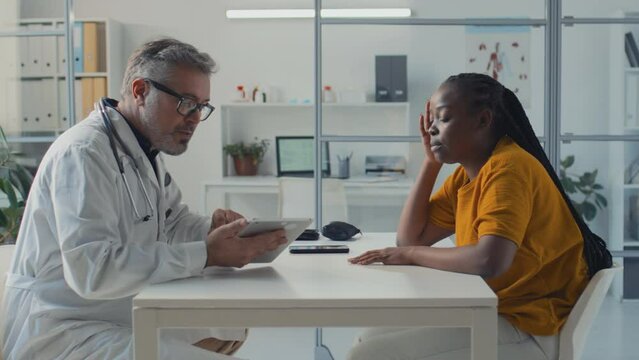 Medium side shot of mature Caucasian male doctor sitting at table with African American female patient, consulting about bloodwork results on tablet computer, and giving therapy recommendations